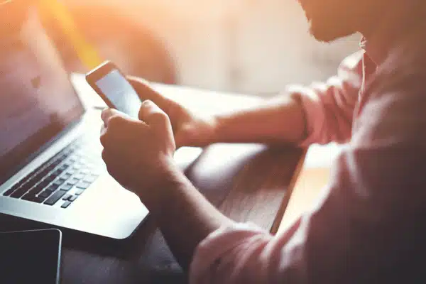 Man checks smartphone beside open laptop, reviewing emails and system notifications in sunlight at desk