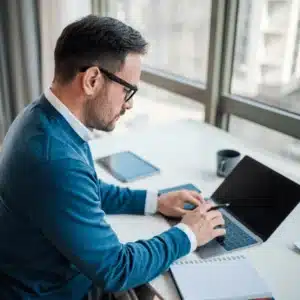Professional man types on a laptop while referencing notes, working remotely by a large window