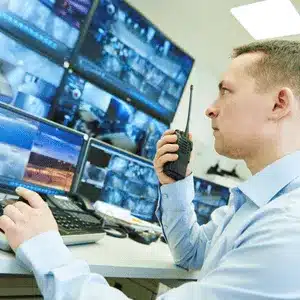 Guard monitors security camera footage across several screens while communicating through a walkie-talkie