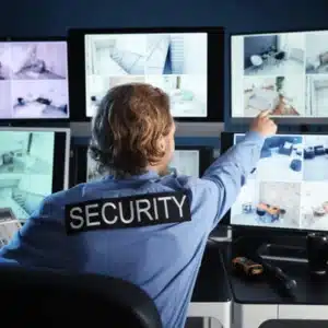 Operator in SECURITY shirt points at monitors, checking security services equipment cameras for suspicious activity