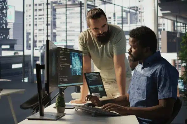 Two men in a modern office discuss data and code displayed on multiple computer screens