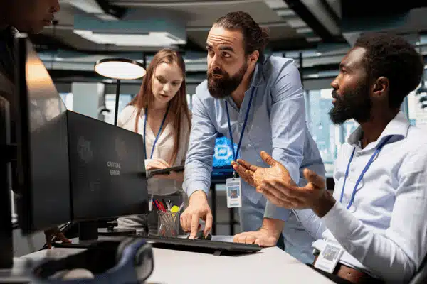 Team of IT professionals discuss critical error on monitor, coordinating response in operations center today