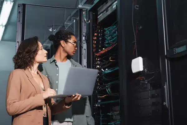 Two network technicians inspect server racks while referencing a laptop; photo of data center interior