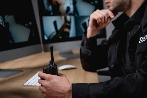 Security officer holds radio, monitors multiple surveillance screens, watching for incidents and coordinating responses tonight
