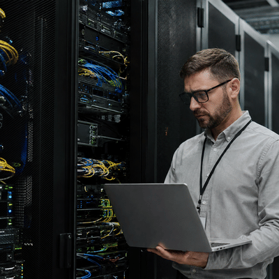 An IT professional reviews rack-mounted systems on a laptop while performing server hardening inside a secure data center.
